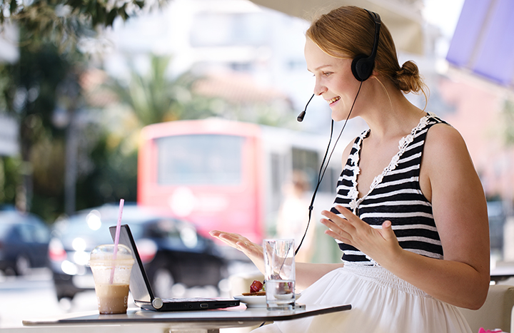 Laughing attractive blond woman wearing a headset sitting with a laptop at an open-air restaurant enjoying refreshments and talking to her friend using skype